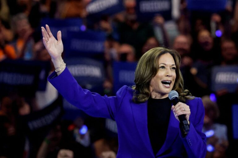 U.S. Vice President Kamala Harris in a blue suit, speaking into a microphone at her rally in Milwaukee, Wisconsin, for a live broadcast to the Democratic National Convention