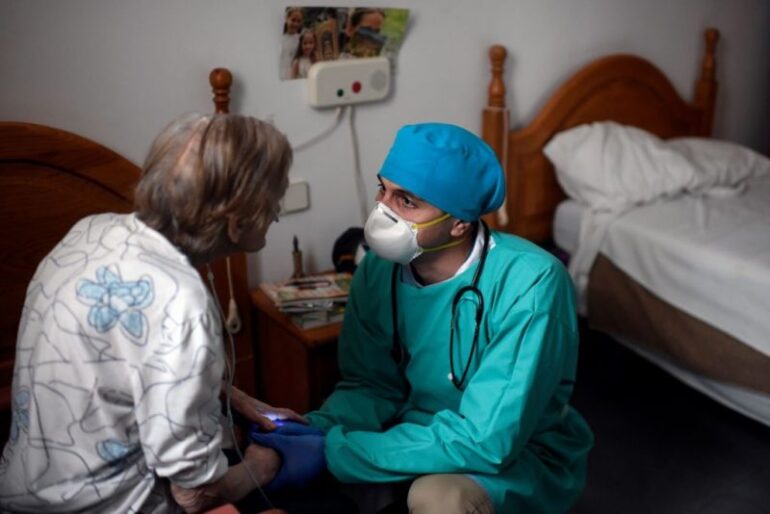 TOPSHOT - Venezuelan doctor Diego Padron holds hands of an isolated resident at an elderly people nursing home in Madrid on April 24, 2020. - What mistakes led to so many deaths? Spain begins to investigate the tragedies behind the closed doors of its retirement homes, where thousands of deaths are attributed to coronavirus. (Photo by OSCAR DEL POZO / AFP) (Photo by OSCAR DEL POZO/AFP via Getty Images)