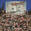 TOPSHOT - Syrian fans cheer for their national team under portraits of late president Hafez al-Assad and current President Bashar al-Assad (R) during their Asian World Cup qualifying football match against Kuwait at the Abbassin Stadium in Damascus on June 2, 2008. Syria won the match 1-0. AFP PHOTO/LOUAI BESHARA (Photo by Louai Beshara / AFP) (Photo by LOUAI BESHARA/AFP via Getty Images)