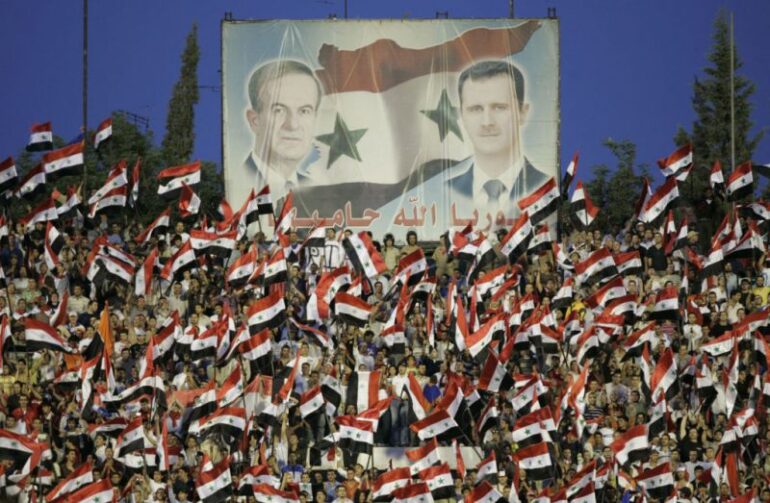 TOPSHOT - Syrian fans cheer for their national team under portraits of late president Hafez al-Assad and current President Bashar al-Assad (R) during their Asian World Cup qualifying football match against Kuwait at the Abbassin Stadium in Damascus on June 2, 2008. Syria won the match 1-0. AFP PHOTO/LOUAI BESHARA (Photo by Louai Beshara / AFP) (Photo by LOUAI BESHARA/AFP via Getty Images)