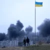 People standing near a Ukrainian national flag watch as dark smoke billows following an air strike in the western Ukrainian city of Lviv, on March 26, 2022. At least five people wounded in two strikes on Lviv, the regional governor said, in a rare attack on a city that has escaped serious fighting since Russian troops invaded last month. (Photo by Oleksii FILIPPOV / AFP) (Photo by OLEKSII FILIPPOV/AFP via Getty Images)