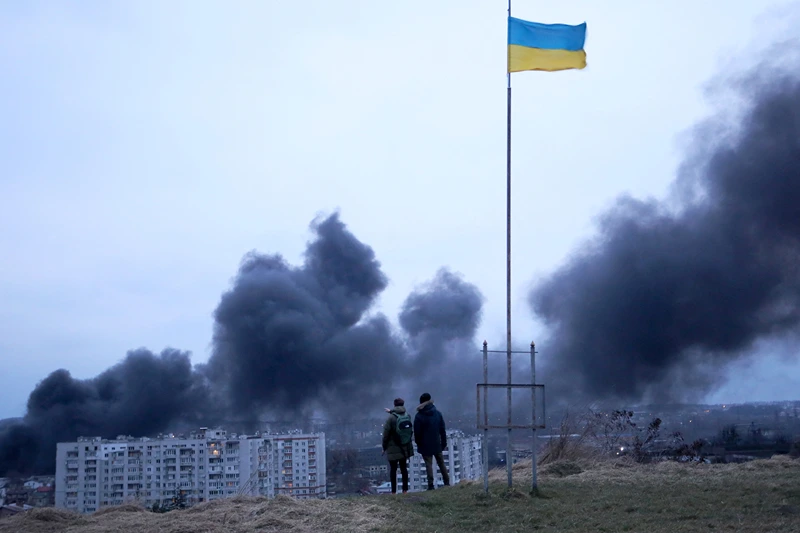 People standing near a Ukrainian national flag watch as dark smoke billows following an air strike in the western Ukrainian city of Lviv, on March 26, 2022. At least five people wounded in two strikes on Lviv, the regional governor said, in a rare attack on a city that has escaped serious fighting since Russian troops invaded last month. (Photo by Oleksii FILIPPOV / AFP) (Photo by OLEKSII FILIPPOV/AFP via Getty Images)