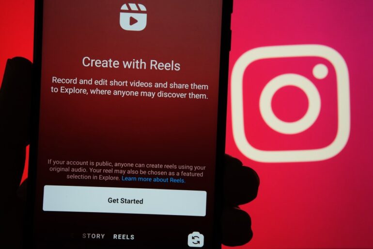 Hand holding a mobile phone displaying the start screen of Instagram Reels feature, with Instagram logo on blurred background.