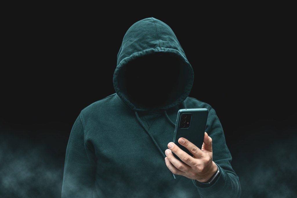 Silhouette of a faceless, hooded figure, presumably a hacker or cybercriminal, holding a smartphone