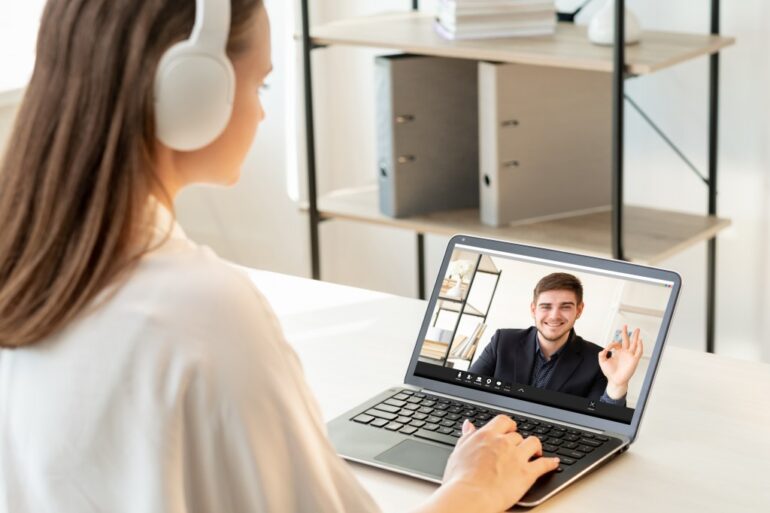 Male teacher giving encouraging OK gesture to female student via video chat on laptop for online education at a modern light workplace