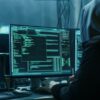 Hooded hacker working on multiple computer screens displaying code and a world map.