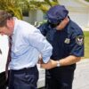 A police officer handcuffing a man in formal attire outside a police car