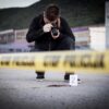 Crime scene photographer behind police tape with evidence marker.