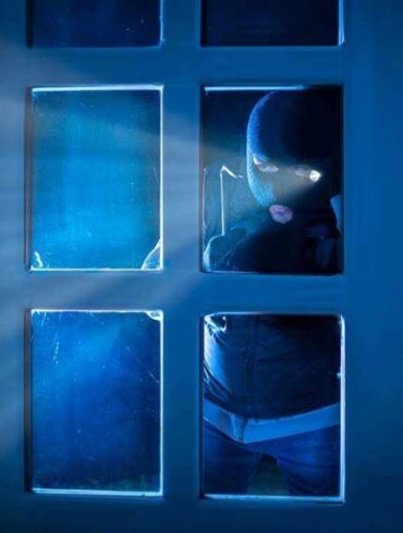 Masked figure peering through glass door at night.