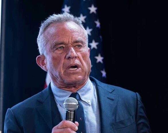 Man holding microphone, speaking with American flag behind.