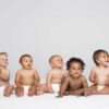 Five diverse babies sitting on a neutral background, displaying playful expressions