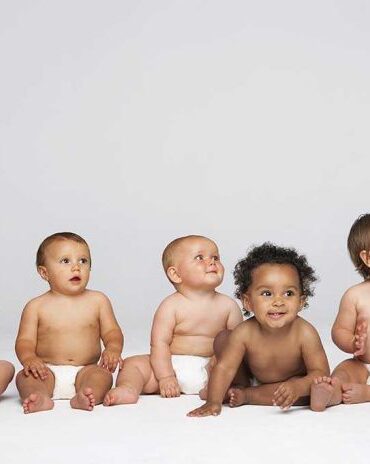 Five diverse babies sitting on a neutral background, displaying playful expressions