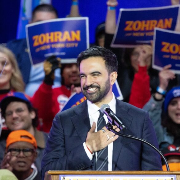Zohran Mamdani at a rally with a mic and his hand over his heart, surrounded by supporters holding "Zohran for New York City" signs.