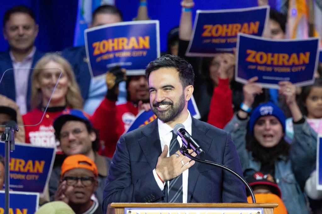 Zohran Mamdani at a rally with a mic and his hand over his heart, surrounded by supporters holding "Zohran for New York City" signs.