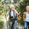 Happy senior couple hiking with trekking sticks and backpacks at the young pine forest