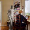 NEO humanoid robot assisting an elderly woman sewing while a man stands in the background.