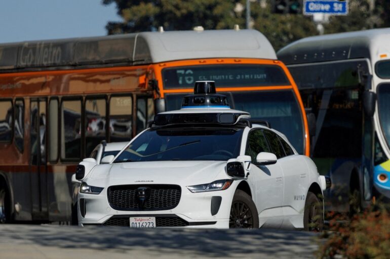 A white Waymo driverless car in traffic in downtown Los Angeles.
