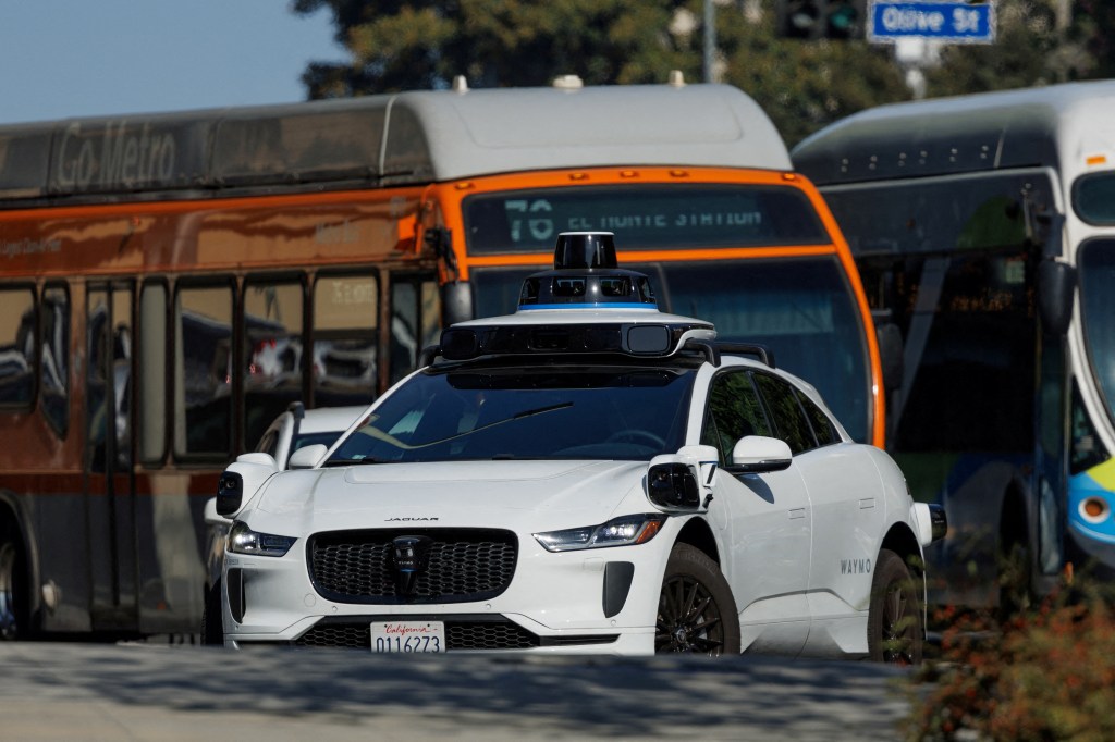 A white Waymo driverless car in traffic in downtown Los Angeles.
