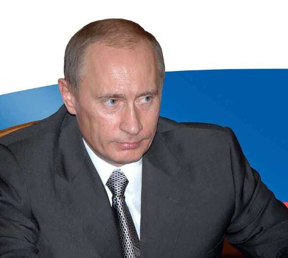 A serious-looking man in a suit sitting at a table with a Russian flag in the background