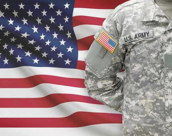 Soldier in camouflage uniform standing before American flag.