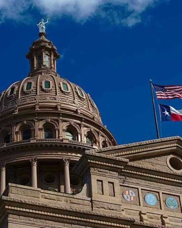 Building dome with US and Texas flags, blue sky.
