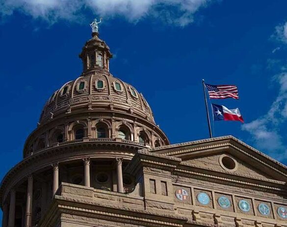 Building dome with US and Texas flags, blue sky.