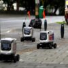 Three delivery robots with orange flags on a paved sidewalk next to a street.