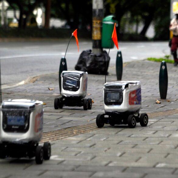 Three delivery robots with orange flags on a paved sidewalk next to a street.