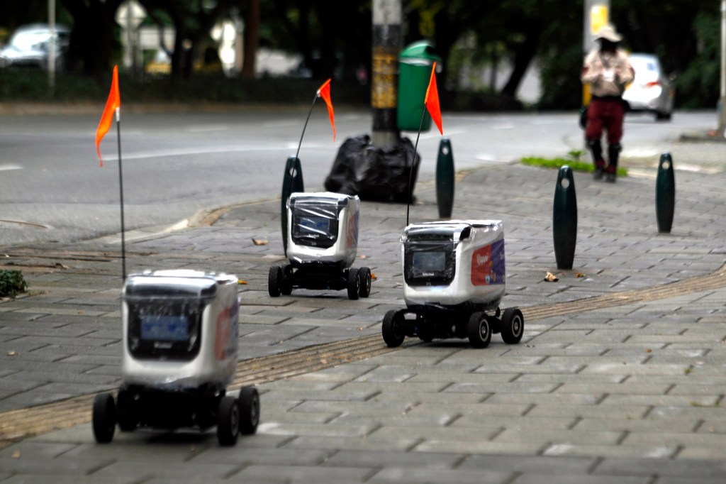 Three delivery robots with orange flags on a paved sidewalk next to a street.