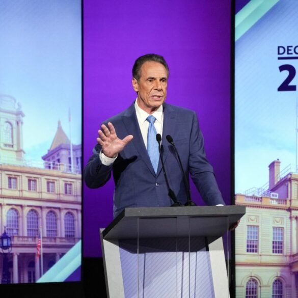 Andrew Cuomo at a debate podium with a large screen displaying "Decision 2025" and a building facade in the background.