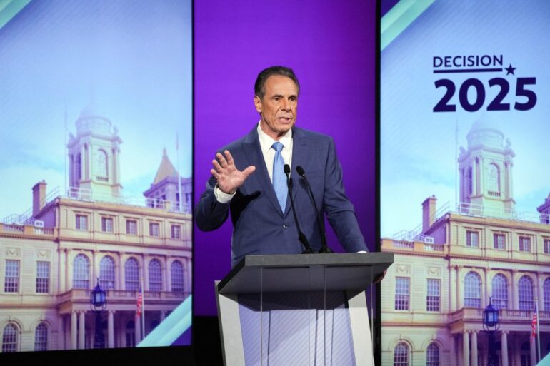Andrew Cuomo at a debate podium with a large screen displaying "Decision 2025" and a building facade in the background.
