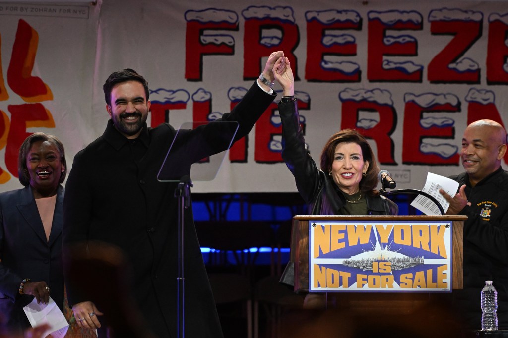 Zohran Mamdani and Kathy Hochul raise joined hands, with other politicians, in front of a "New York is Not For Sale" banner.