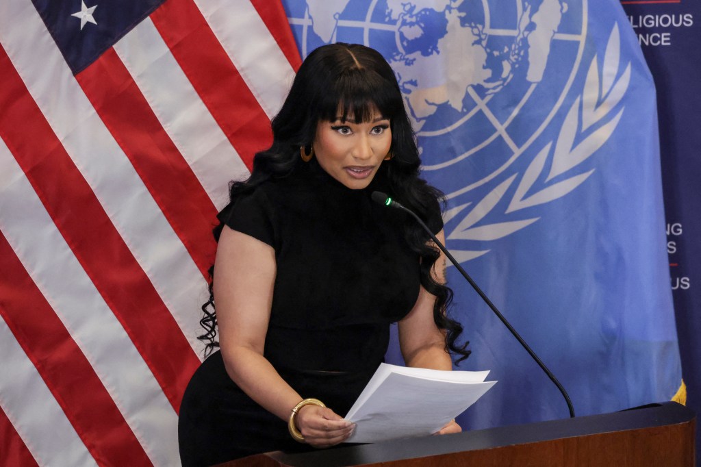 Nicki Minaj speaking at a podium with a microphone, holding papers, in front of US and UN flags.