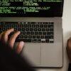 A hacker typing on a MacBook laptop with code on the screen.