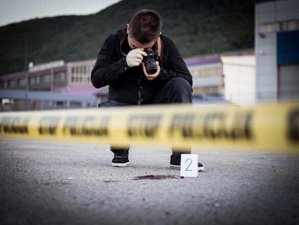 Crime scene photographer behind police tape with evidence marker.