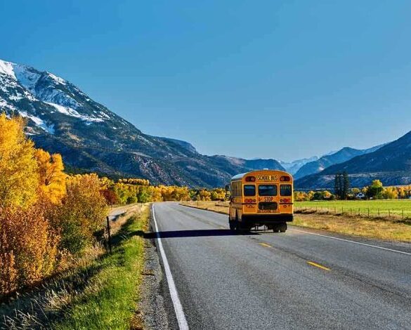 A yellow school bus driving on a rural road surrounded by autumn trees and mountains