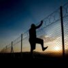 Silhouette of person climbing barbed wire fence at sunset.
