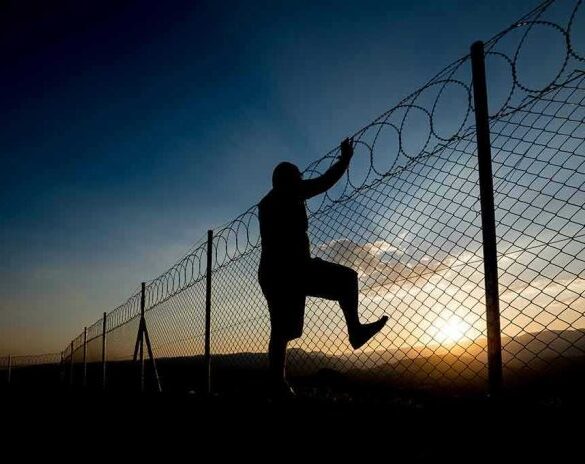 Silhouette of person climbing barbed wire fence at sunset.