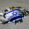 An Israeli flag laid on the ground amongst flowers, tributes, and a small notebook with pens.