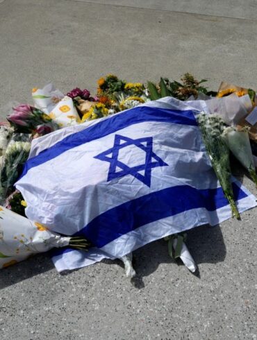 An Israeli flag laid on the ground amongst flowers, tributes, and a small notebook with pens.