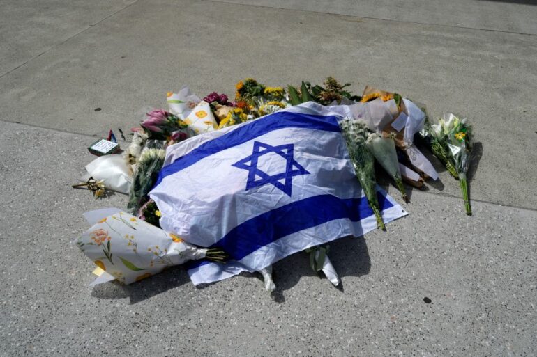An Israeli flag laid on the ground amongst flowers, tributes, and a small notebook with pens.