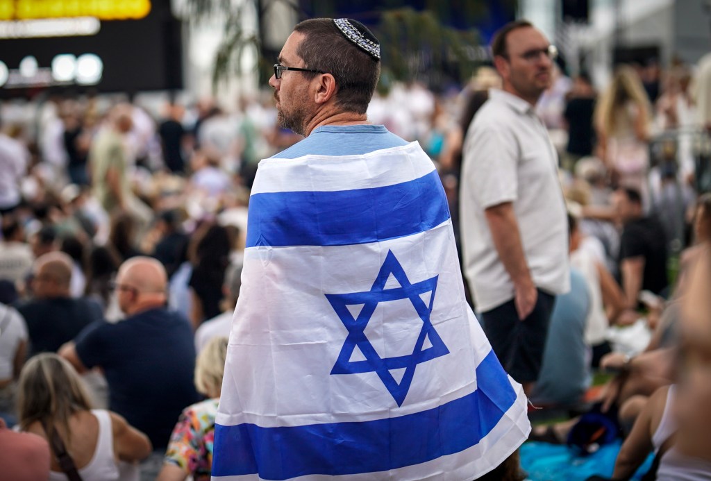 A man wearing a kippah and an Israeli flag around his shoulders attends a memorial vigil.