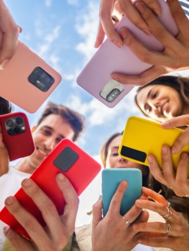 A low-angle photo of a diverse group of young people, smiling and standing in a circle, looking at their colorful mobile phones.