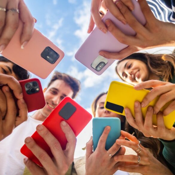 A low-angle photo of a diverse group of young people, smiling and standing in a circle, looking at their colorful mobile phones.