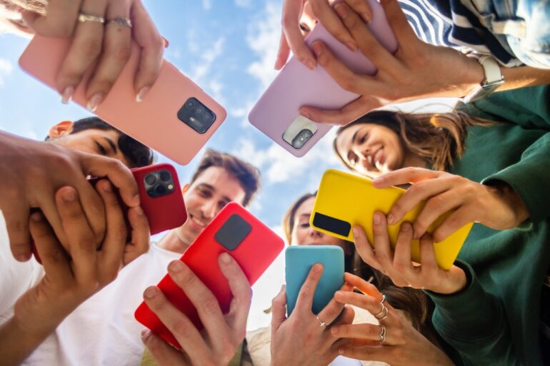 A low-angle photo of a diverse group of young people, smiling and standing in a circle, looking at their colorful mobile phones.