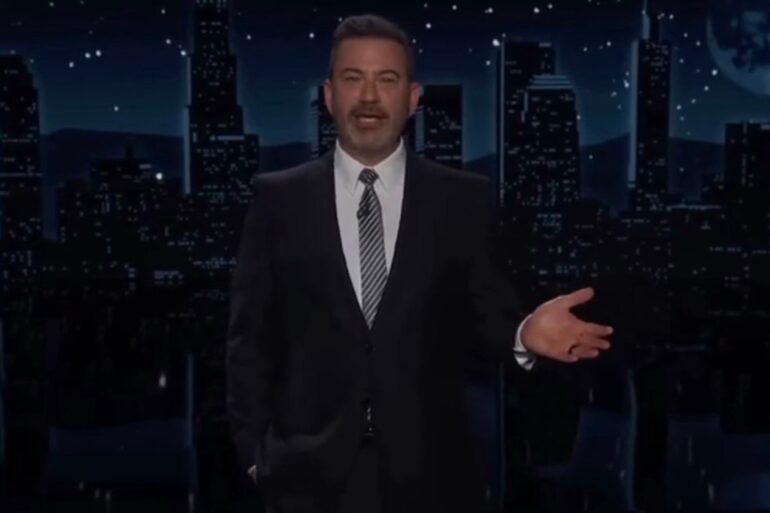 Jimmy Kimmel standing in front of a city skyline at night, gesturing with his right hand.