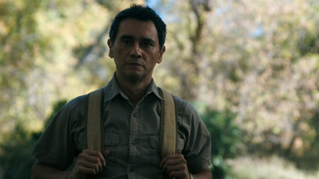 Manousos holding onto his backpack straps in the jungle in Pluribus episode 7