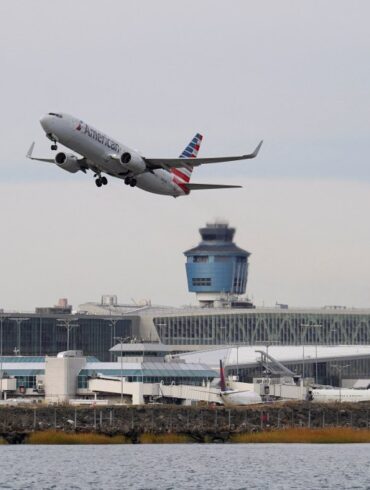 An American Airlines plane takes off from LaGuardia Airport over a body of water, with the air traffic control tower and airport terminals visible.