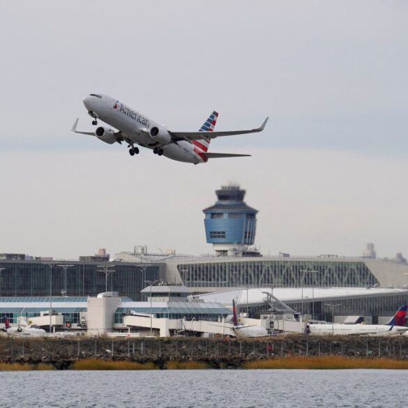 An American Airlines plane takes off from LaGuardia Airport over a body of water, with the air traffic control tower and airport terminals visible.