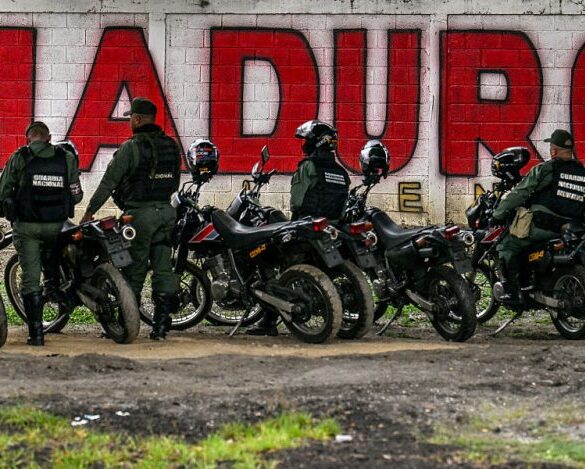Members of the National Guard stand guard in front of a mural of ousted Venezuela's President Nicolas Maduro in Caracas on January 10, 2026. The US State Department on January 10, urged Americans in Venezuela to leave the country "immediately," citing risks from armed militias searching vehicles for US citizens at roadblocks. (Photo by Juan BARRETO / AFP via Getty Images)